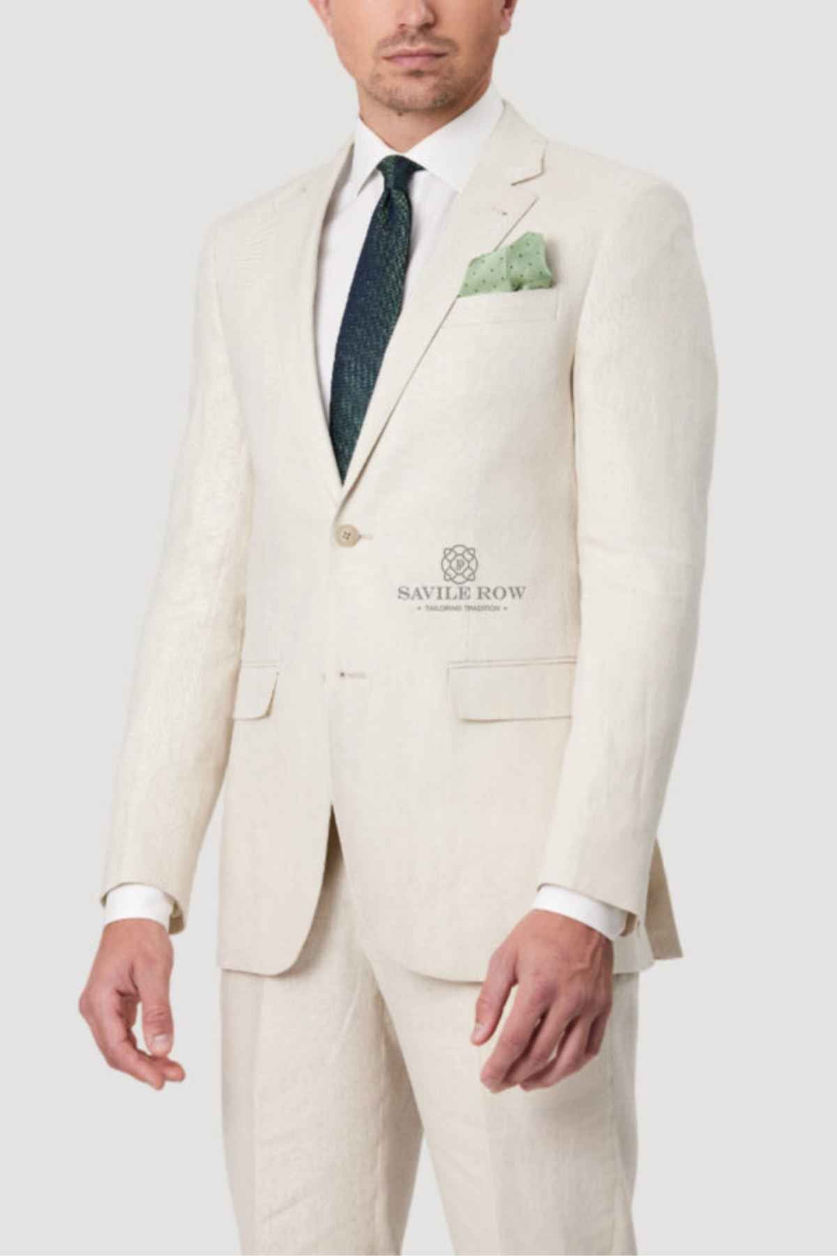 A man dressed in the Bruton Abram Pure Linen Blazer in ecru, paired with a white shirt, dark green tie, and light green pocket square, stands against a plain light gray backdrop. The blazer is lightweight with two buttons and features two front pockets.