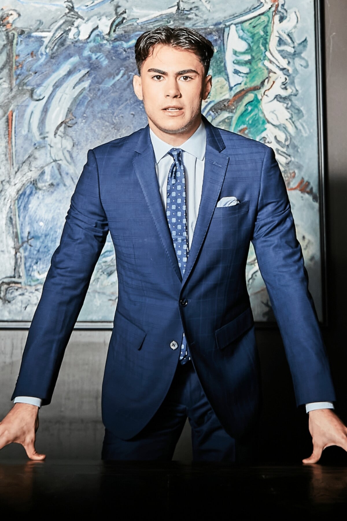 Dressed in a Bruton Abram Check Suit Jacket, the man stands confidently with hands on a table in front of an abstract painting, embodying formal elegance.