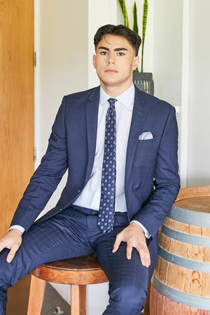 Wearing a Bruton Abram Check Suit Jacket and tie, a man sits on a wooden stool next to a barrel. The room features a tall plant and wooden door, creating an ambiance ideal for formal occasions.