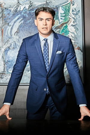 Dressed in a Bruton Abram Check Suit Jacket, the man stands confidently with hands on a table in front of an abstract painting, embodying formal elegance.