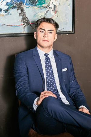 A young man in a Bruton Abram Check Suit Jacket sits with legs crossed. His short dark hair enhances his confident forward gaze. An abstract art piece on the dark wall behind provides an elegant backdrop suited for any formal occasion.