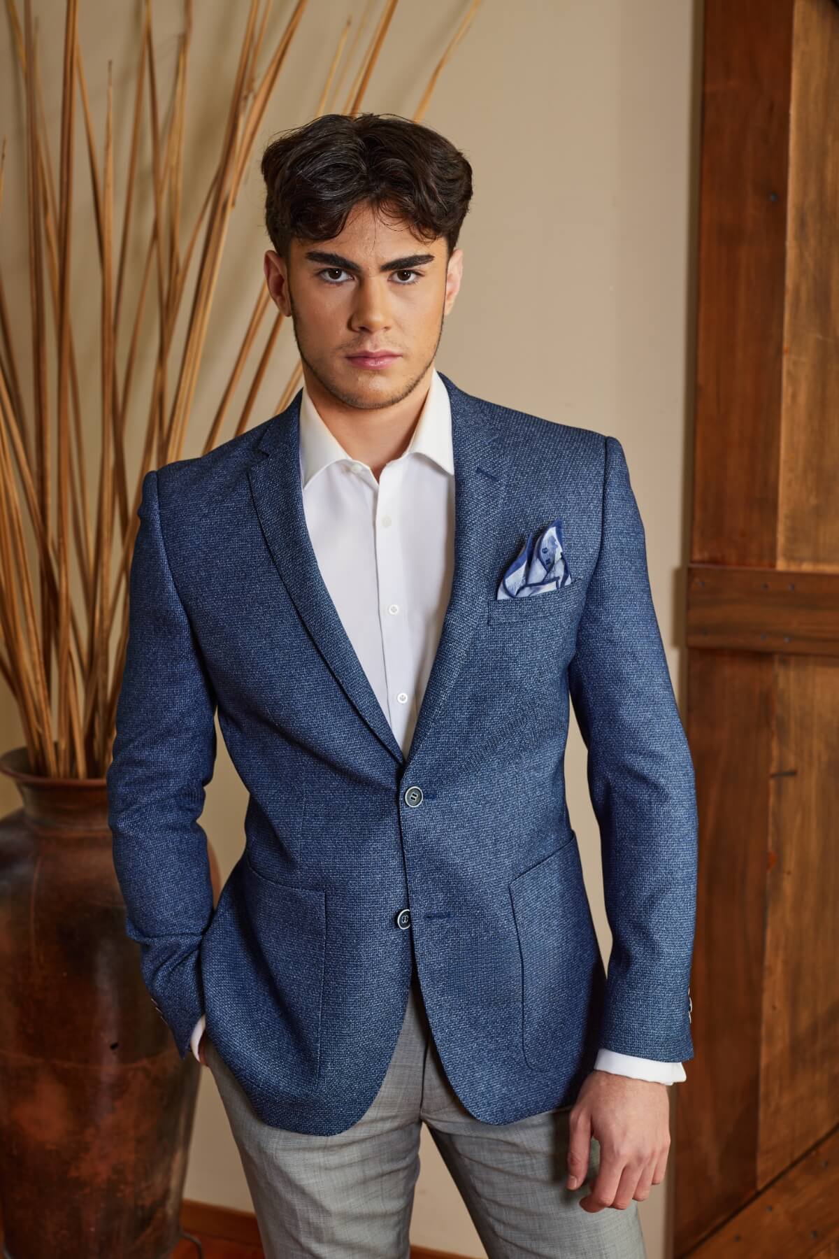 A person with dark hair is wearing a blue Bruton Adin Blazer, white shirt, patterned pocket square, and gray pants. One hand is in their pocket as they stand indoors against a wooden wall, with a decorative vase in the background.