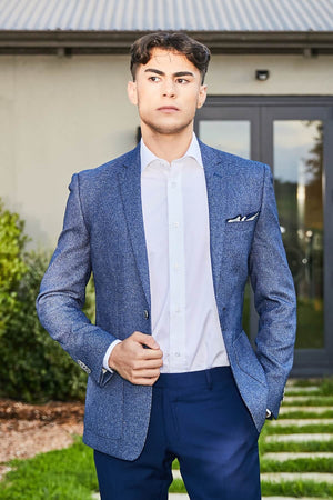 Standing outdoors wearing a Bruton Adin Blazer, white shirt, and dark pants, the person features a slim fit blazer with a pocket square and geometric texture. Slightly looking off-camera, there's a backdrop of buildings, greenery, and pathways.
