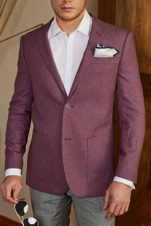 A man dressed in a white dress shirt, a red Bruton Agar Blazer accessorized with a pocket square, and gray pants is holding a pair of sunglasses in his right hand. The modern fit of the blazer complements his tailored look perfectly against the backdrop of a wooden surface adorned with some abstract wall art.