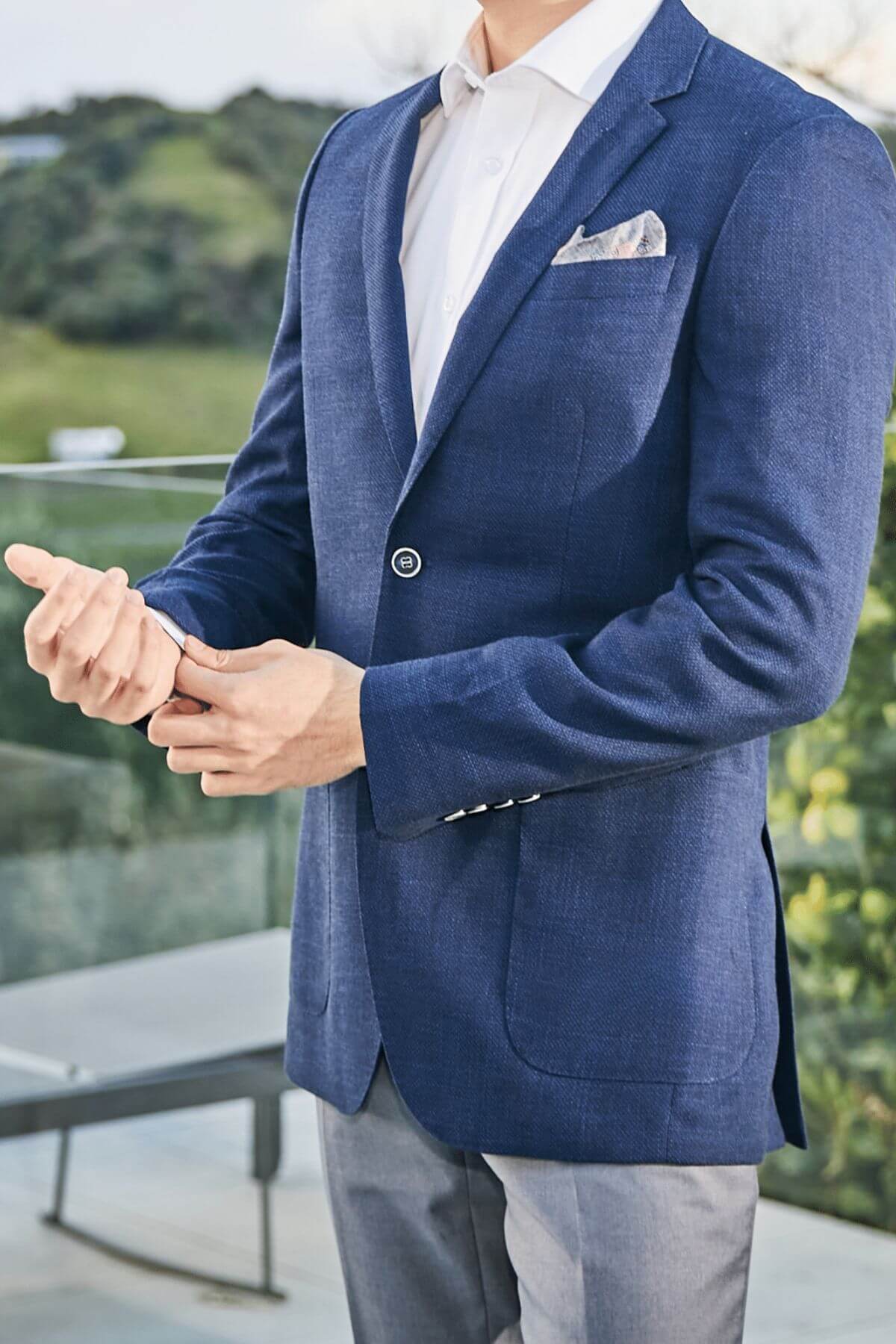 Wearing the Bruton Agar Blazer, a man in a modern fit with reinforced shoulder pads, white shirt, and gray pants stands outdoors on a patio, adjusting his sleeve with greenery and a glass railing behind him.