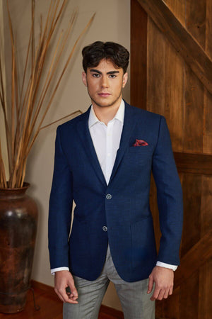 A young man with dark hair and a serious expression stands indoors near a large clay vase and wooden door, wearing the Bruton Agar Blazer over a white dress shirt and gray trousers.