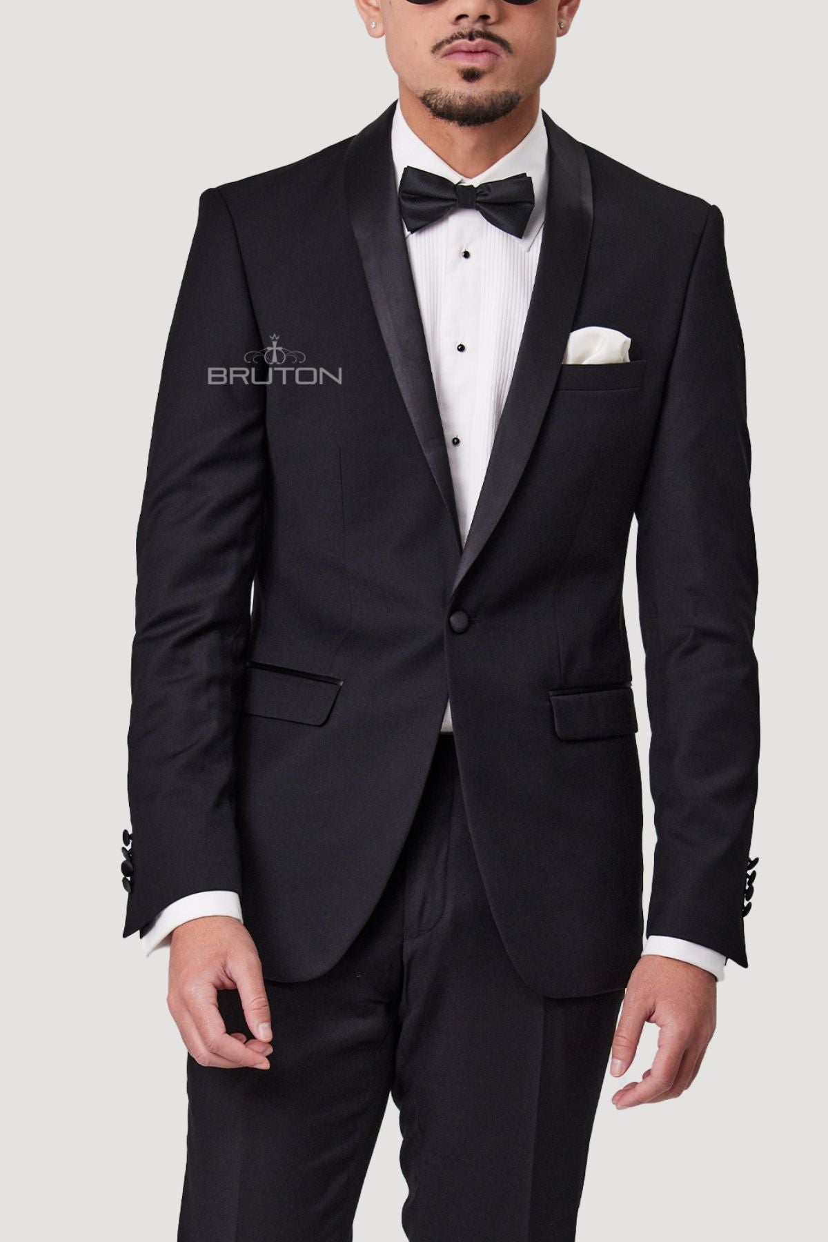 A man in a slim-fit tuxedo, crafted from SSA8 BLK fabric, with satin lapels stands against a plain background. Sporting short hair and facial hair, he completes the look with a white dress shirt, black bow tie, and a pocket square accenting his Bruton Benhur Shawl Collar Jacket.