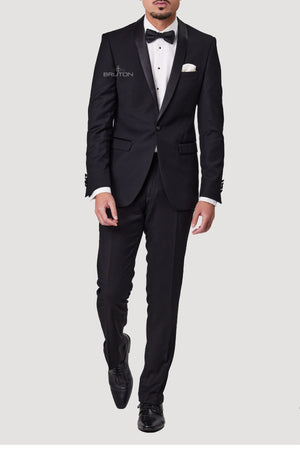 A man in a slim-fit tuxedo featuring a Bruton Benhur Shawl Collar Jacket crafted from SSA8 BLK fabric with satin lapels and a logo on the chest. He wears a white dress shirt, black bow tie, and matching dress shoes. His right hand rests in his pocket, adorned with a crisp white pocket square.