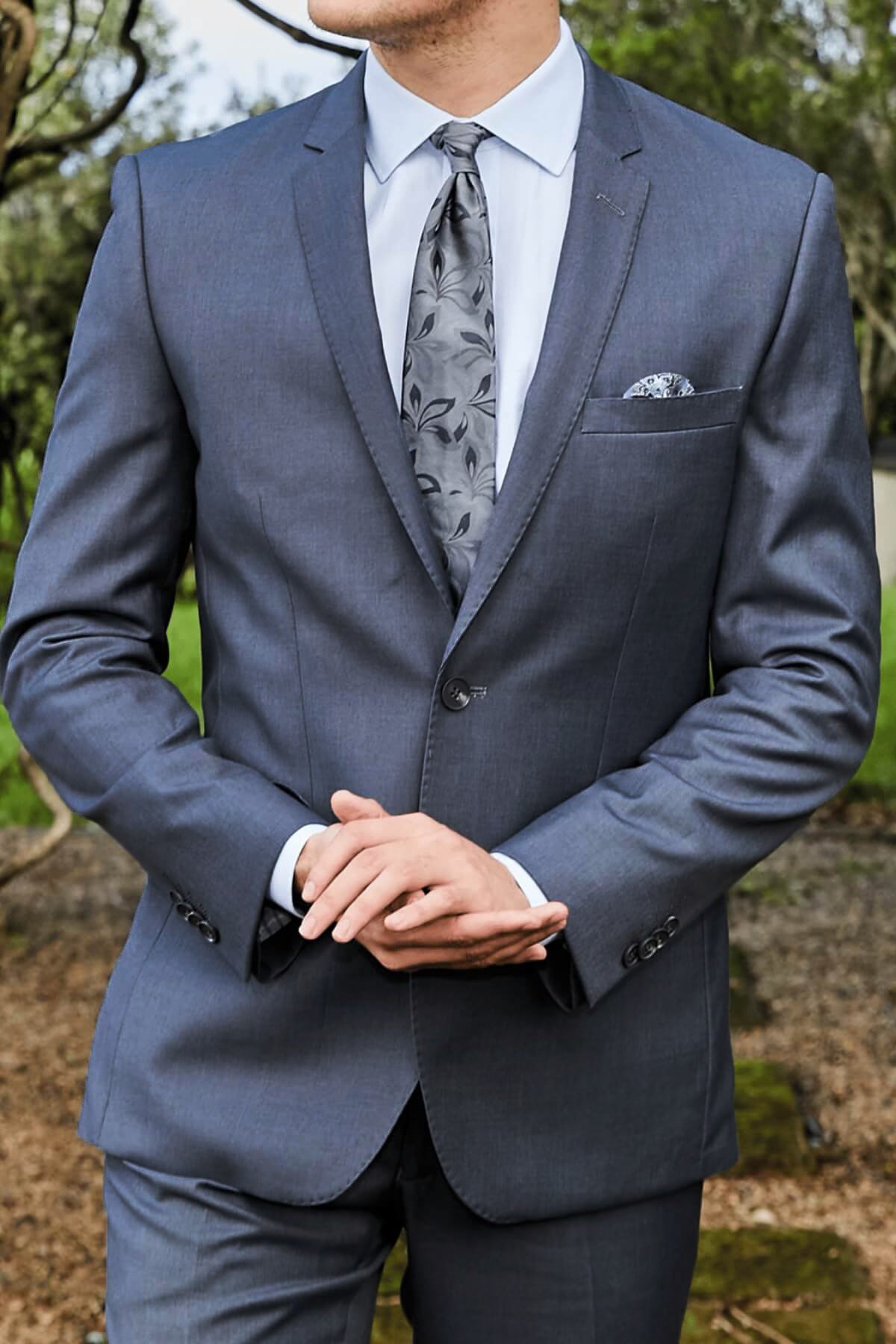 A man outdoors, hands clasped, wears the Bruton David Suit Jacket with a matching patterned tie and pocket square; its slim fit and structured shoulders stand out against greenery.
