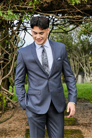 A young man wears the Bruton David Suit Jacket and a patterned tie, smiling outdoors with one hand in his pocket against a backdrop of leafy greenery.