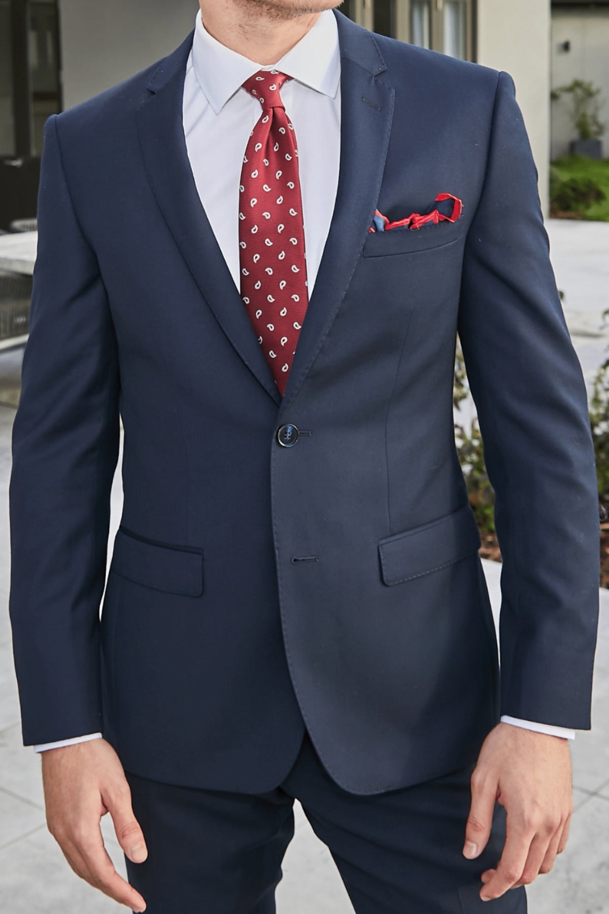 A man, partially out of frame, stands outdoors wearing the Bruton David Suit Jacket in slim fit dark blue with structured shoulders, paired with a white shirt, red patterned tie, and matching pocket square.