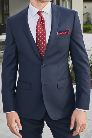 A man, partially out of frame, stands outdoors wearing the Bruton David Suit Jacket in slim fit dark blue with structured shoulders, paired with a white shirt, red patterned tie, and matching pocket square.