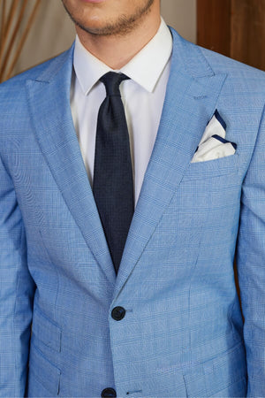 A person in a slim fit light blue checkered suit with a white shirt and dark tie, wearing the Bruton Jed Suit Jacket featuring a peak lapel and a neatly placed white pocket square. The background is softly blurred.