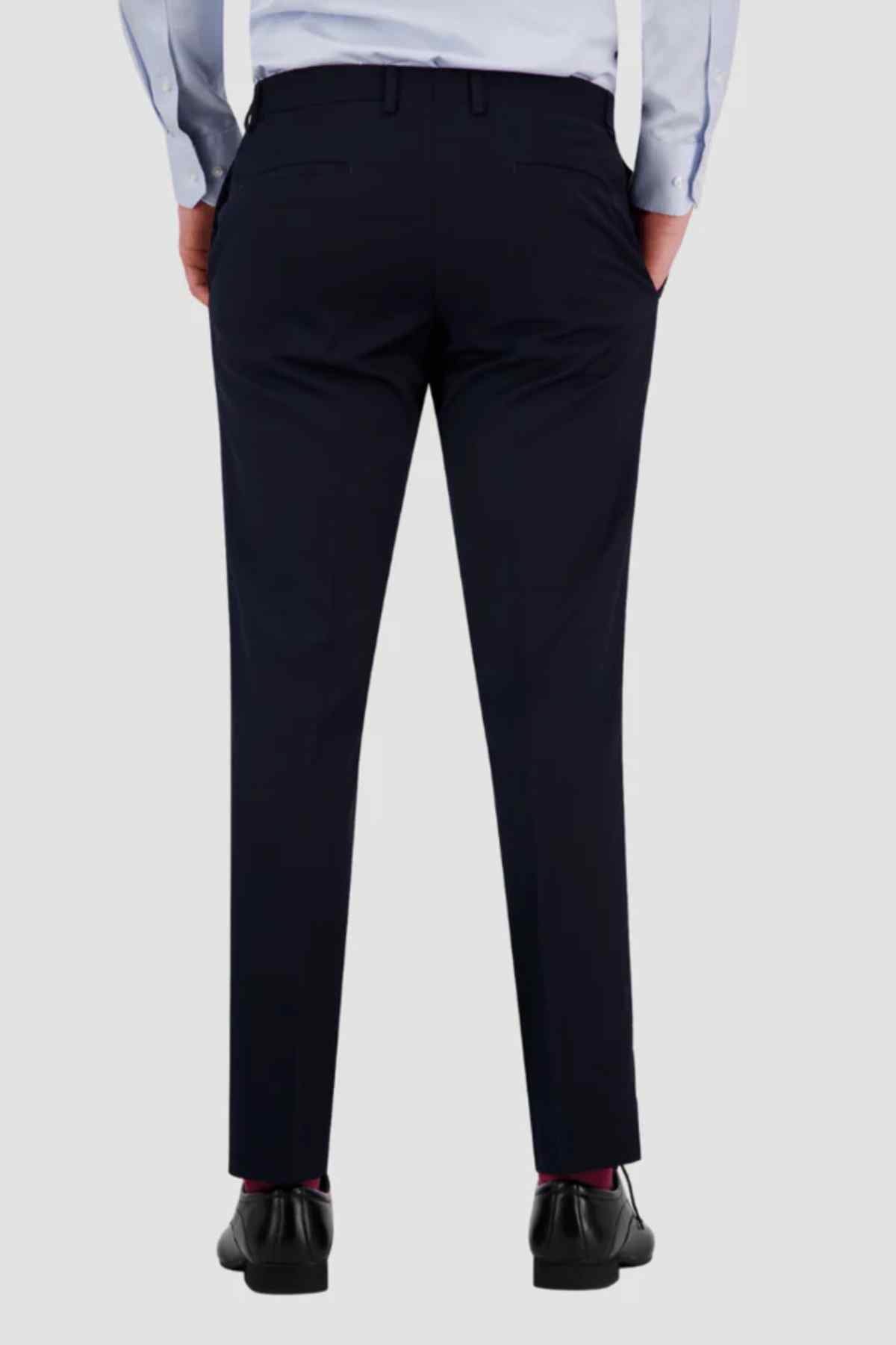 Rear view of a person wearing Bruton Jesse Trouser in navy blue slim fit with buttoned back pockets, paired with a light blue shirt and black dress shoes, standing hands-in-pockets against a plain light background.