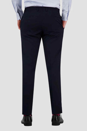 Rear view of a person wearing Bruton Jesse Trouser in navy blue slim fit with buttoned back pockets, paired with a light blue shirt and black dress shoes, standing hands-in-pockets against a plain light background.