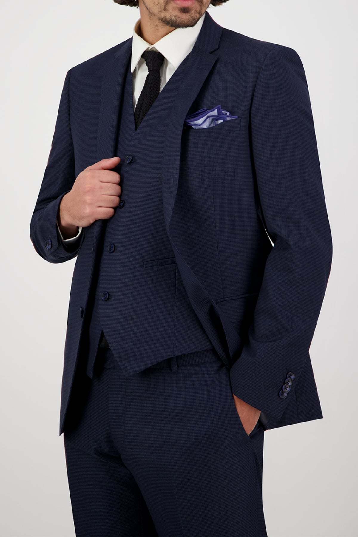 A man in a slim-fit Bruton Jose Suit Jacket (dark blue) pairs it with a white dress shirt, black tie, and purple pocket square. He poses with one hand on his jacket for a sharp look; his face is partially out of frame.