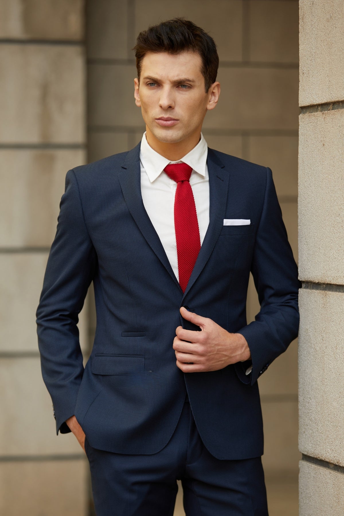 Wearing the slim fit Bruton Jose Suit Jacket with a white shirt and red tie, a man stands confidently against a stone wall, glancing to the side while adjusting his jacket for a sharp, polished appearance.