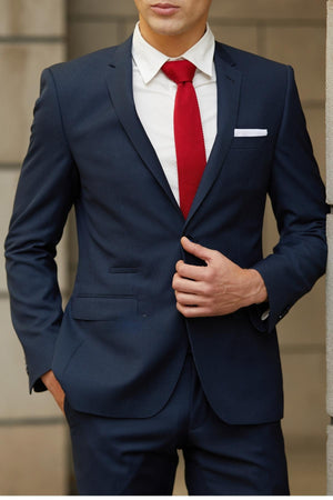 A man in a Bruton Jose Suit Jacket, paired with a white shirt, red tie, and white pocket square, stands with one hand in his pocket and the other adjusting his jacket. His face is partly visible against a blurred background.