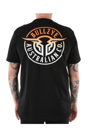 A tattooed person wears the Bullzye Bullring Tee, a black cotton jersey featuring a bold "BULLZYE AUSTRALIAN CO." back print in white and orange. The rear-view photo showcases the tee’s standout design.