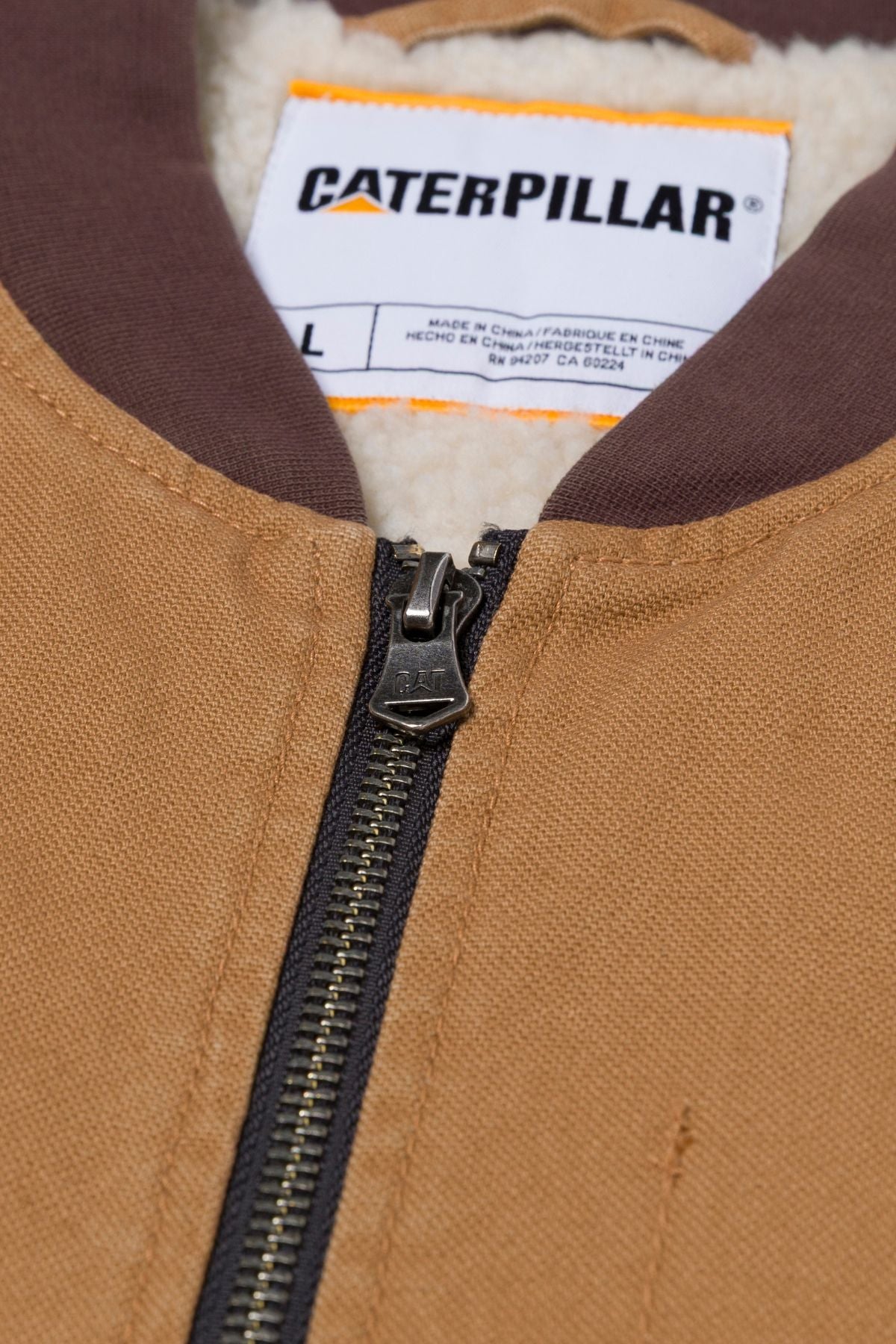 Close-up of a tan CAT Canvas Utility Sherpa Lined Vest with a brown collar, visible zipper, white CAT logo label (size L), and sherpa fleece lining showing near the collar.