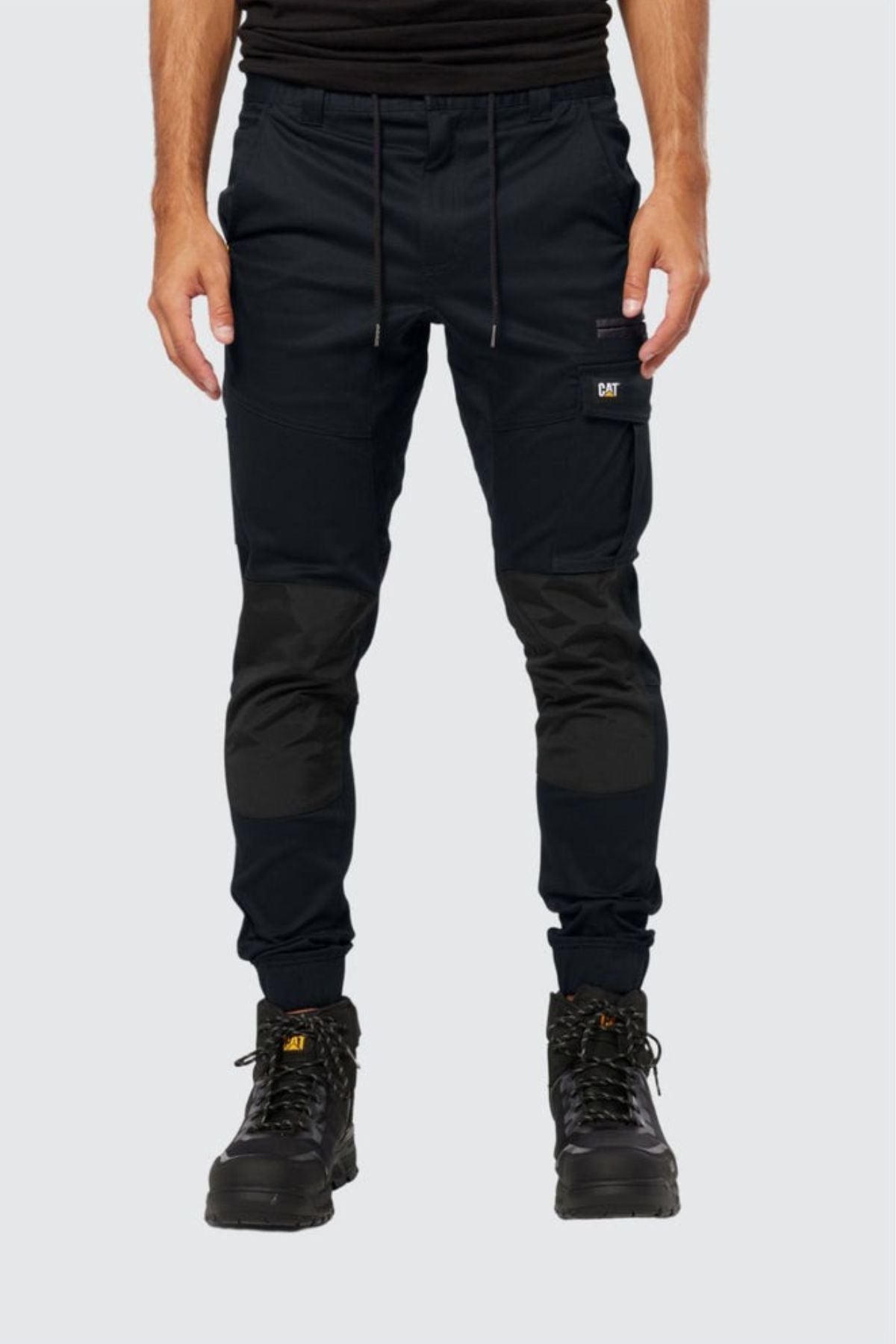 A person in black CAT Cuffed Dynamic Pant—durable work pants with reinforced knees—black CAT boots, and a black shirt, stands against a plain light grey background.