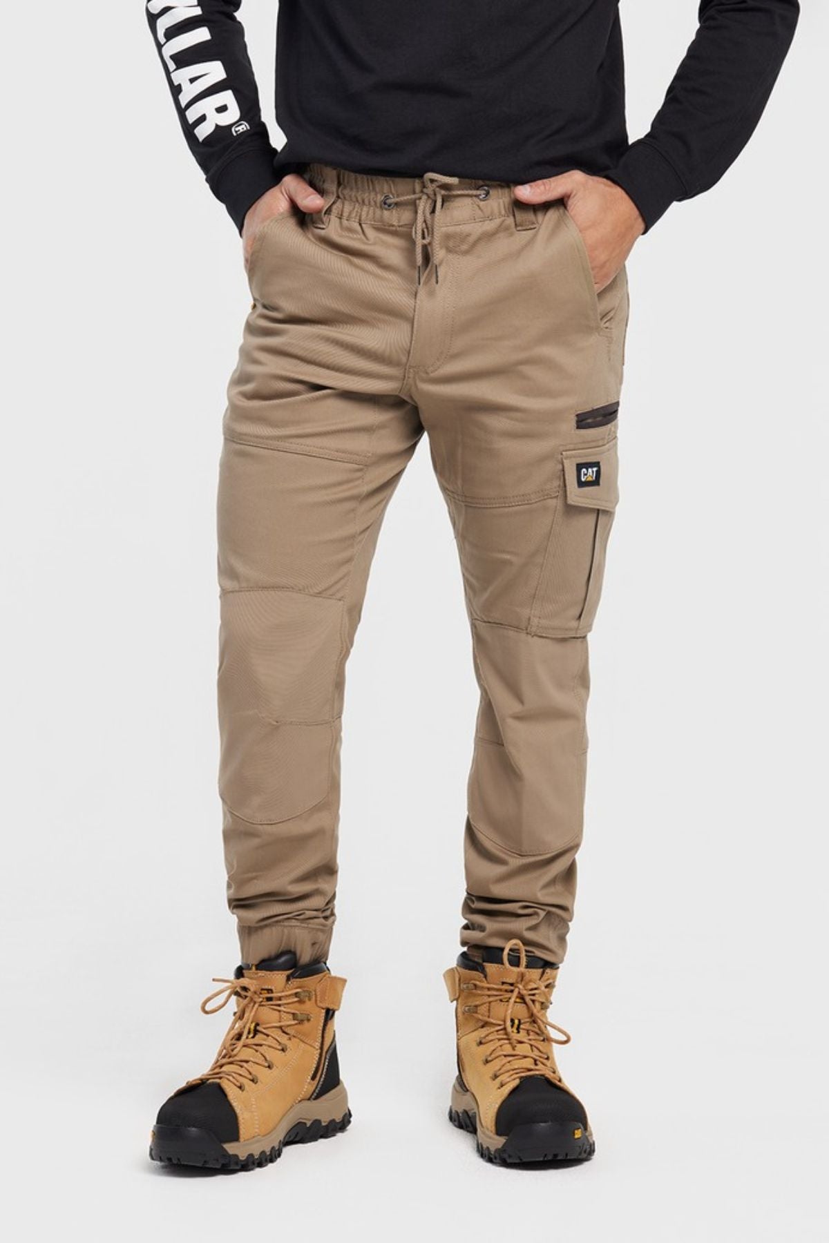 A person stands against a plain white background, wearing tan CAT Cuffed Dynamic Pant joggers, a black long-sleeve shirt, and tan work boots, with hands in pockets.