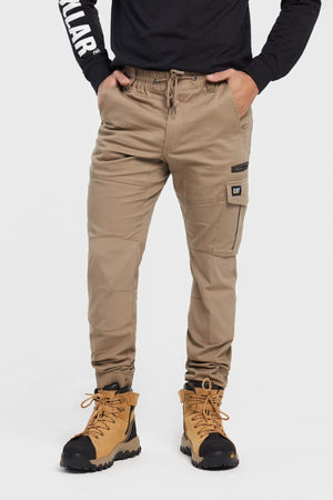A person stands against a plain white background, wearing tan CAT Cuffed Dynamic Pant joggers, a black long-sleeve shirt, and tan work boots, with hands in pockets.