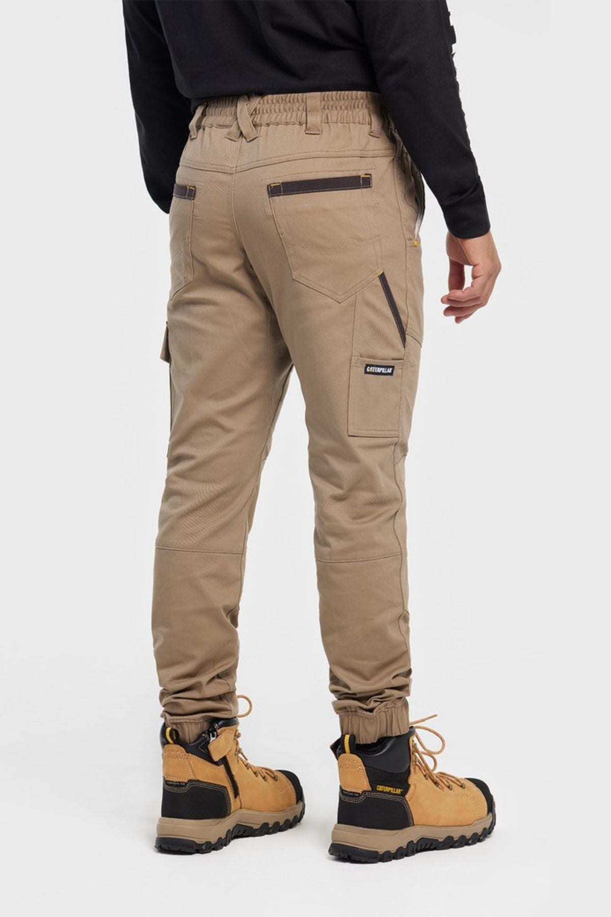 A person faces away in CAT Cuffed Dynamic Pant with zippered back pockets, a side pocket label, and brown work boots featuring black soles and laces, all set against a plain white background.