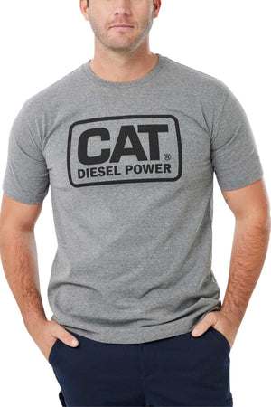 A man with short hair wears a gray CAT Diesel Power Tee featuring black text on the front and a rib collar for style, standing with his hands in his navy blue pants pockets.