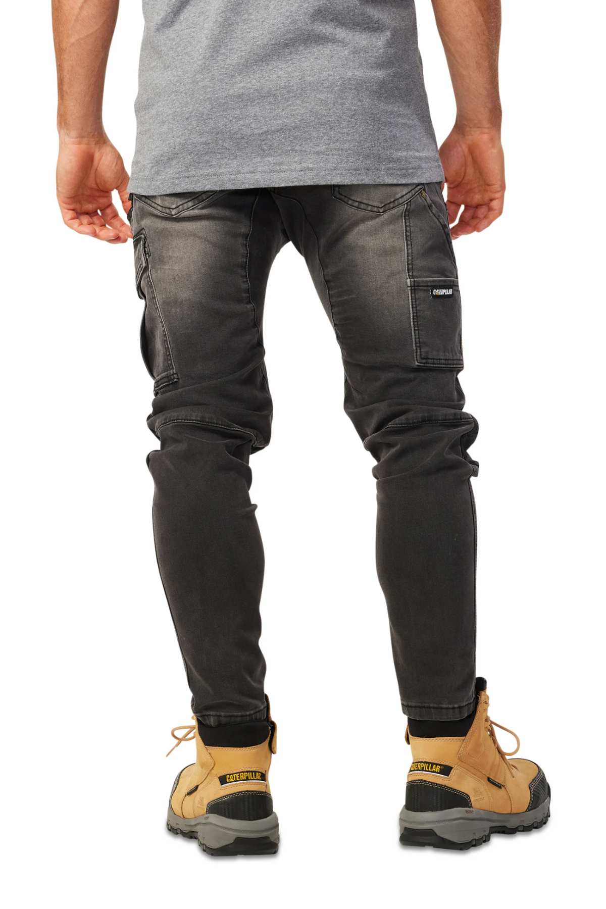 A person is shown from the back wearing dark gray CAT Dynamic Denim Pants with multiple pockets and stitching details, paired with tan work boots and a gray shirt.