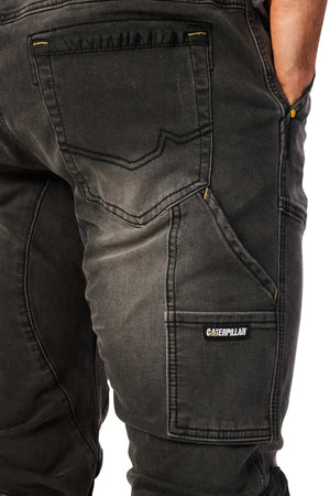 A close-up of a person wearing the stylish CAT Dynamic Denim Pant, showcasing dark, robust fabric with clear stitching and pockets. The Caterpillar logo stands out on a side pocket as the person casually rests their hand in another pocket.