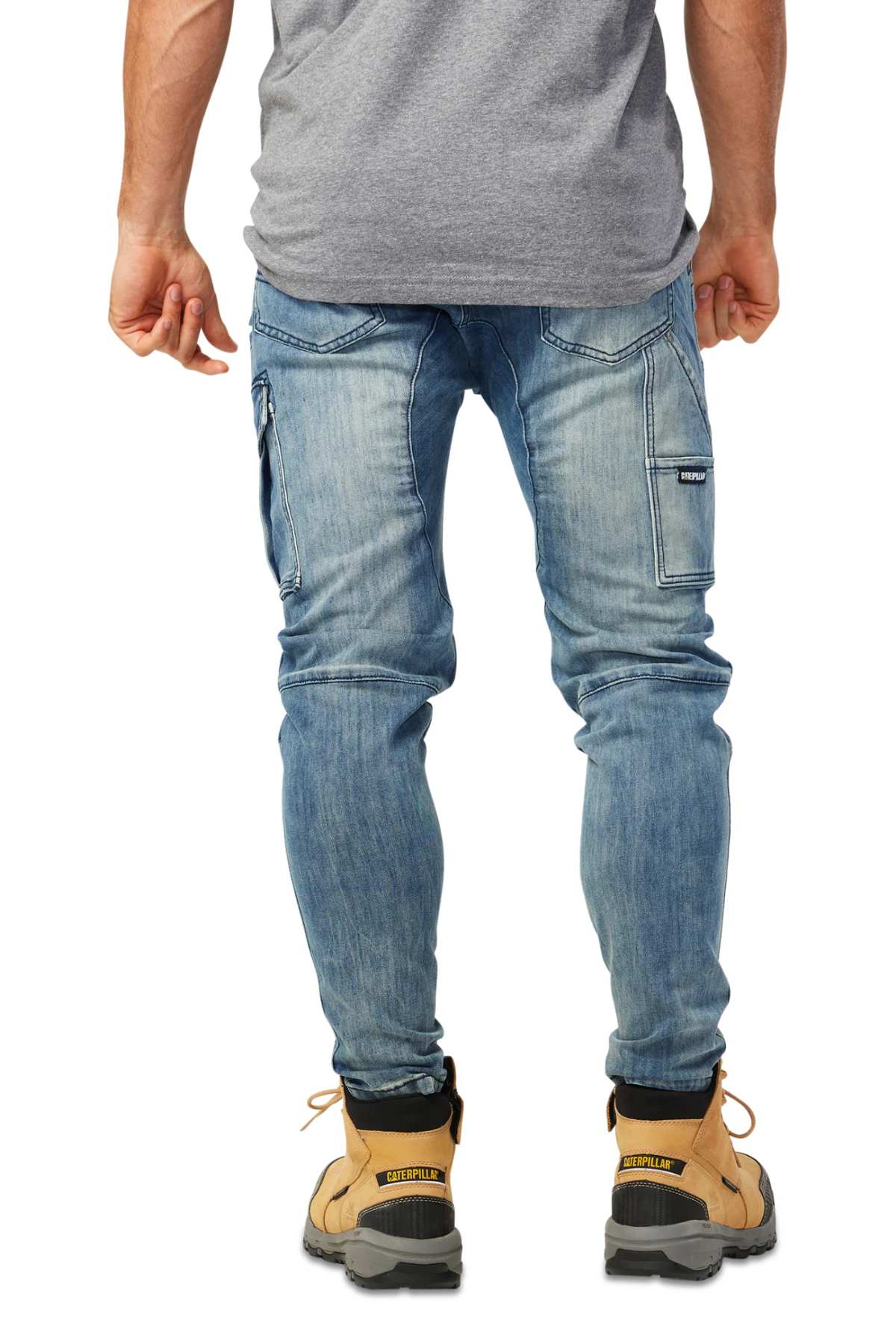 A man wearing CAT Dynamic Denim Pant in light blue with a cargo side pocket, a gray T-shirt, and tan work boots stands with his back to the camera. The fitted jeans offer added utility with their distinctive side pocket.