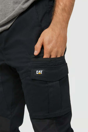 A person wearing stylish black CAT Dynamic Slim Fit Pants, featuring reinforced knee patches and a hand resting on a cargo pocket; only the lower torso and upper legs are visible.