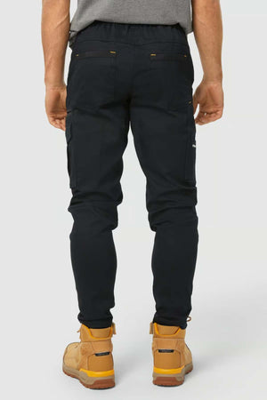 A person wearing the CAT Dynamic Slim Fit Pant in black, paired with a gray shirt and tan work boots, stands facing away from the camera against a plain light background.