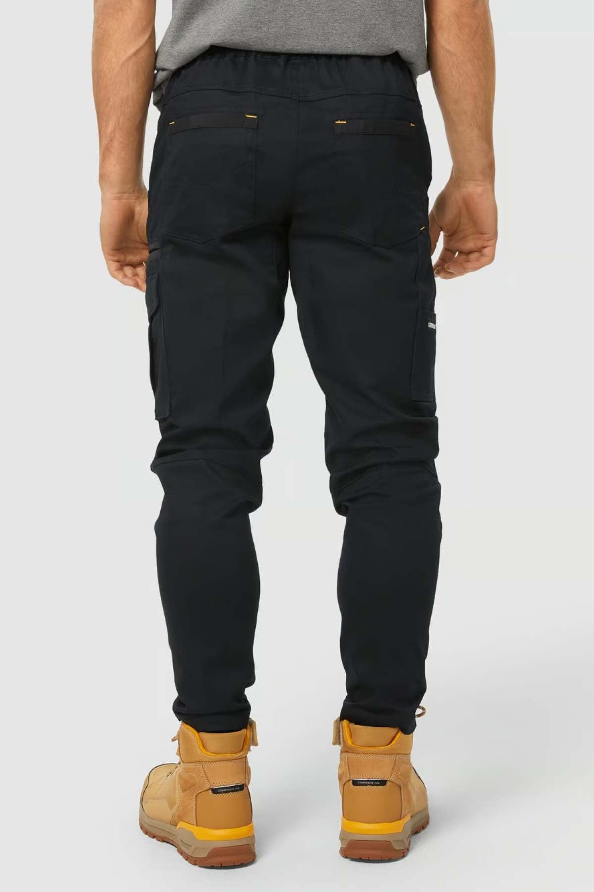 A person is shown from the back wearing black CAT Dynamic Slim Fit Pants with reinforced knee patches and multiple pockets, along with tan work boots, against a plain light background. Their hands are relaxed at their sides.