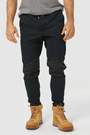 A person wears the CAT Dynamic Slim Fit Pant in black, featuring reinforced knee patches, paired with a gray shirt and tan lace-up work boots. The photo is cropped from the shoulders down against a plain light background.
