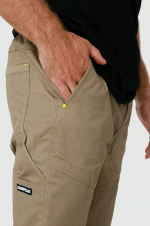 A person stands with their right hand in the pocket of khaki CAT Dynamic Pants featuring a Caterpillar logo on the cargo pocket, paired with a black shirt.
