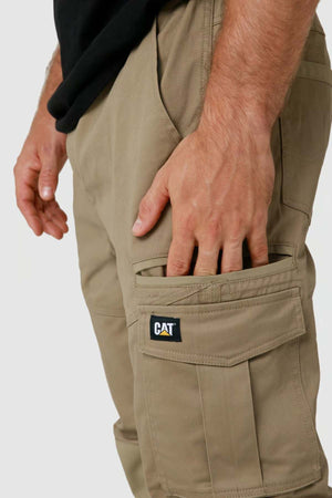 A person dons the beige "CAT Dynamic Slim Fit Pant" with a black "CAT" logo on the cargo pocket, featuring reinforced knee patches. Their hand rests casually on the side, and they wear a partially visible black shirt.