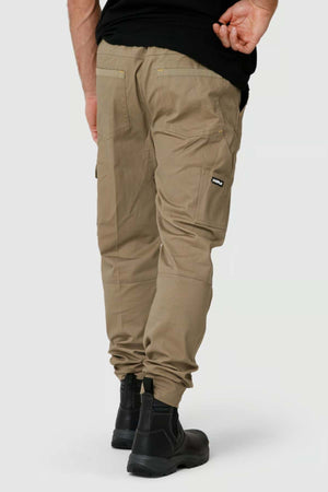 Someone wears durable tan CAT Dynamic Pants with reinforced knees, a black shirt, and black boots while facing away, showcasing the back pockets and cargo pocket details against a light gray background.