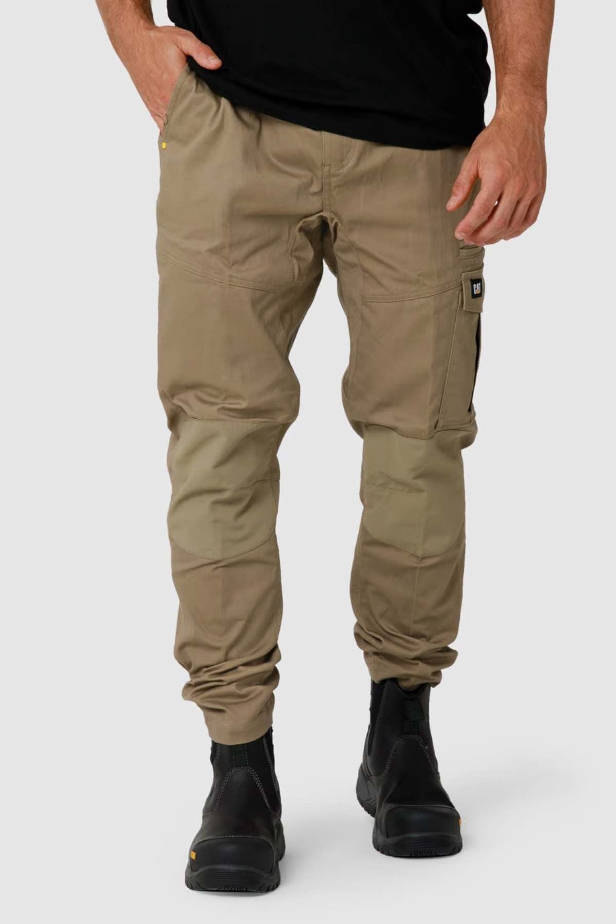 Wearing CAT Dynamic Slim Fit Pants with reinforced knee patches and multiple cargo pockets, paired with black work boots, a person stands against a light gray background. The pants complement the black top as their hands rest casually in the pockets.