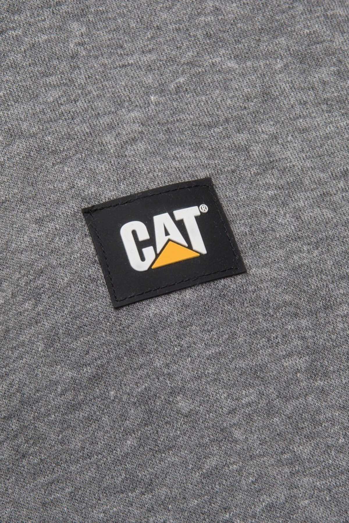 A close-up image of the gray fabric on the CAT Essential 1/4 Zip Sweatshirt shows a black rectangular patch sewn on. The patch features the word "CAT" in bold white letters with a yellow triangle under the letter "A." The sweatshirt boasts super soft fleece lining and nylon overlays at the shoulders.
