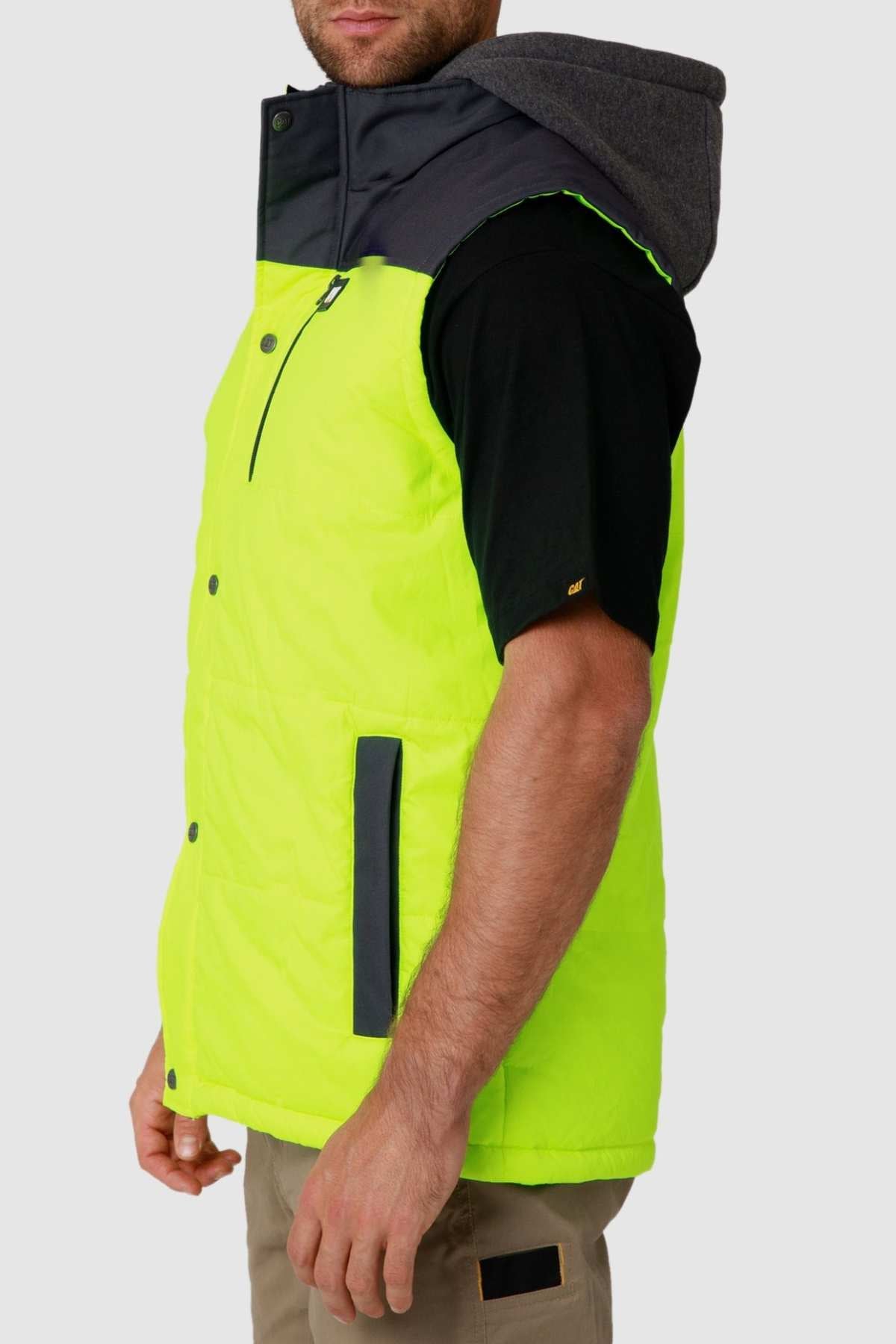 Wearing the CAT Hi Vis Hooded Work Vest, vibrant yellow with black buttons and pockets, over a black t-shirt and paired with tan pants creates a stylish look. The vest is zipped halfway, set against a plain gray background for a blend of functionality and flair.