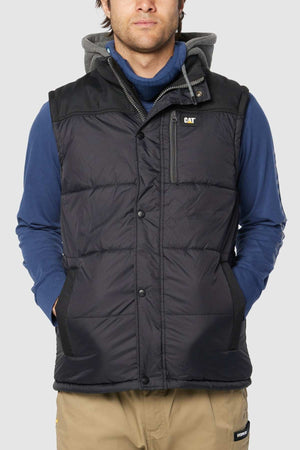 A person is wearing a black CAT Hooded Work Vest, which is water-resistant, over a blue hoodie and beige pants. The vest includes a front zip and button closure with multiple pockets, including a small one on the left chest displaying the "CAT" logo. The person has their hands in the vest pockets and stands against a plain background.