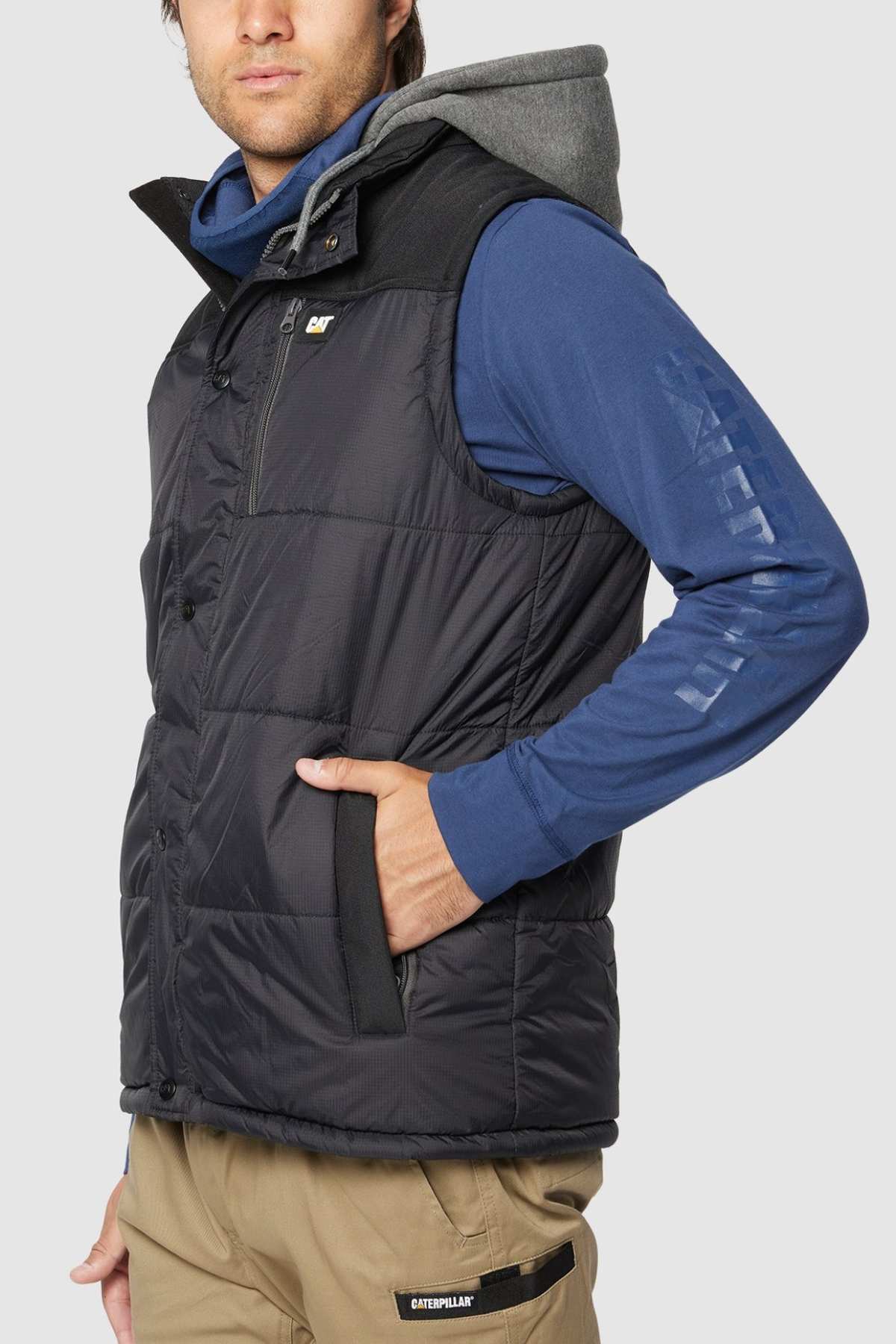 A person is wearing a black CAT Hooded Work Vest, which is water-resistant, over a blue long-sleeve hooded shirt. With their left hand tucked into one of the vest's multiple pockets, they are looking slightly to the left. The lower half of their outfit includes beige pants featuring a visible Caterpillar logo on the pocket.