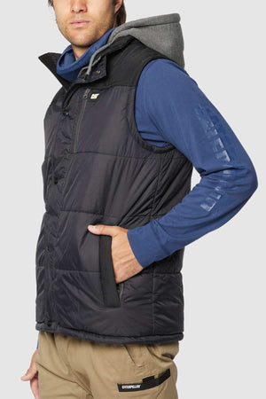 A person is wearing a black CAT Hooded Work Vest, which is water-resistant, over a blue long-sleeve hooded shirt. With their left hand tucked into one of the vest's multiple pockets, they are looking slightly to the left. The lower half of their outfit includes beige pants featuring a visible Caterpillar logo on the pocket.