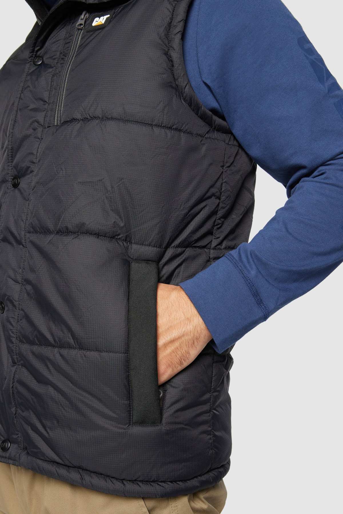A person wearing a CAT Hooded Work Vest, which features multiple pockets, a blue long-sleeve shirt, and beige pants. The person's left hand is tucked into the pocket of the water-resistant vest, highlighting its detailed stitching and zipper. The backdrop is plain and light-colored.