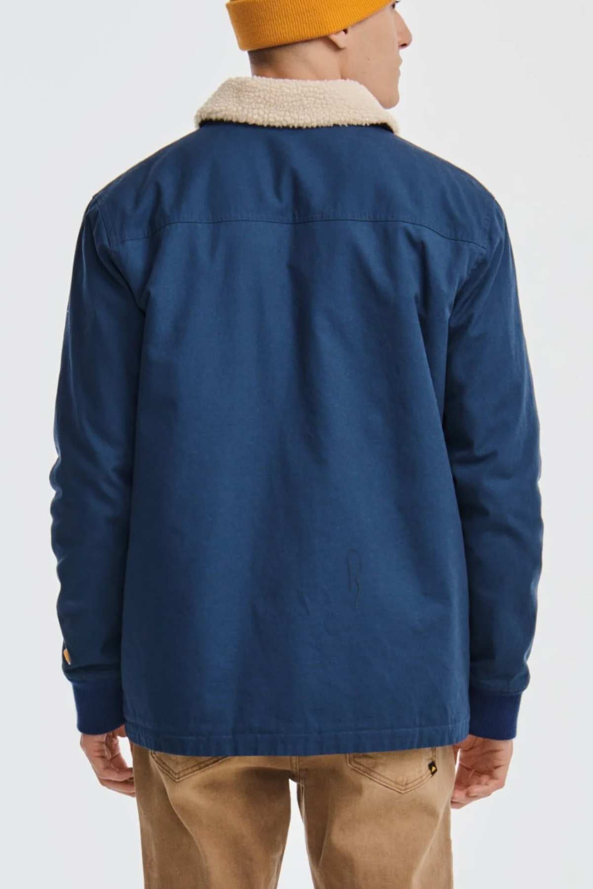 Dressed in a blue CAT Sherpa Lined Chore Jacket featuring a white sherpa collar, an orange beanie, and beige pants, an individual poses against a plain white backdrop.