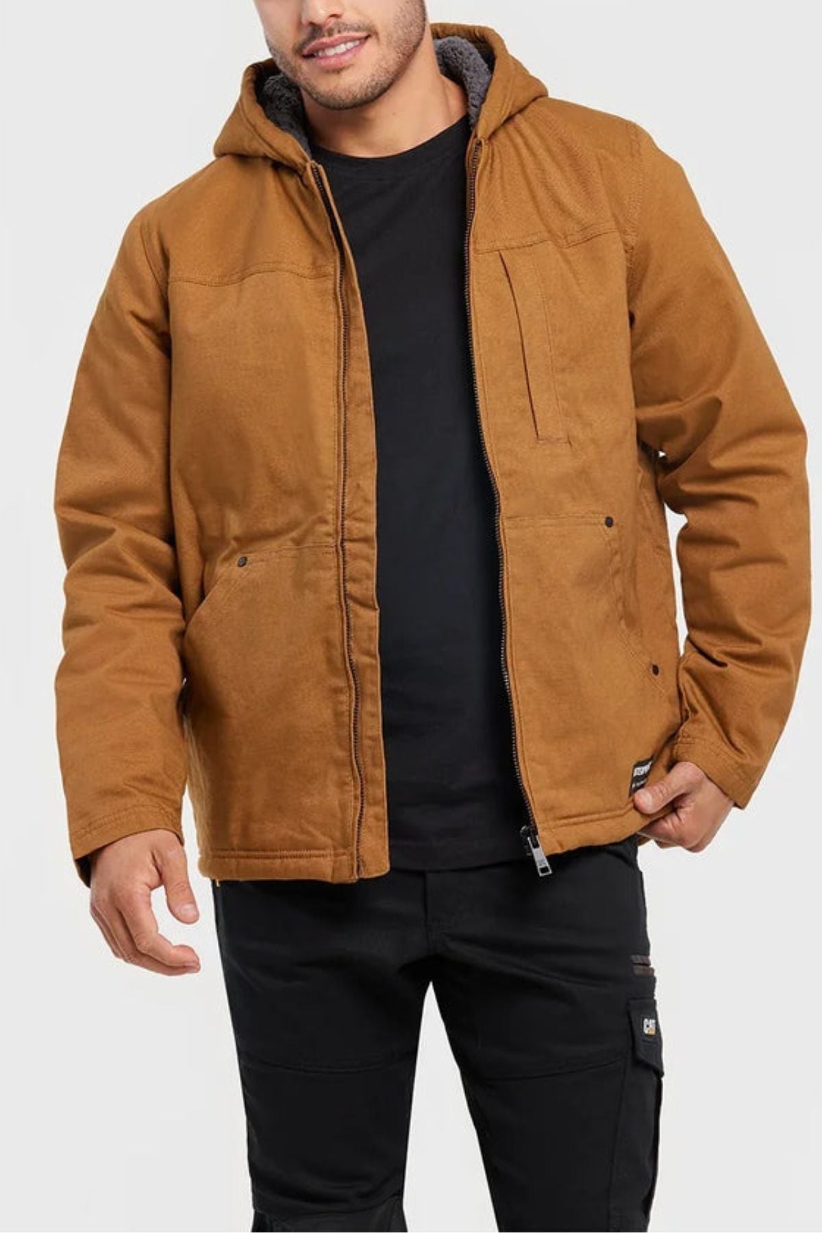 A man stands against a plain light gray background wearing a brown CAT Hooded Sherpa Lined Jacket over a black shirt and black pants. The outdoor jacket features zip and snap closures, plus visible pockets.