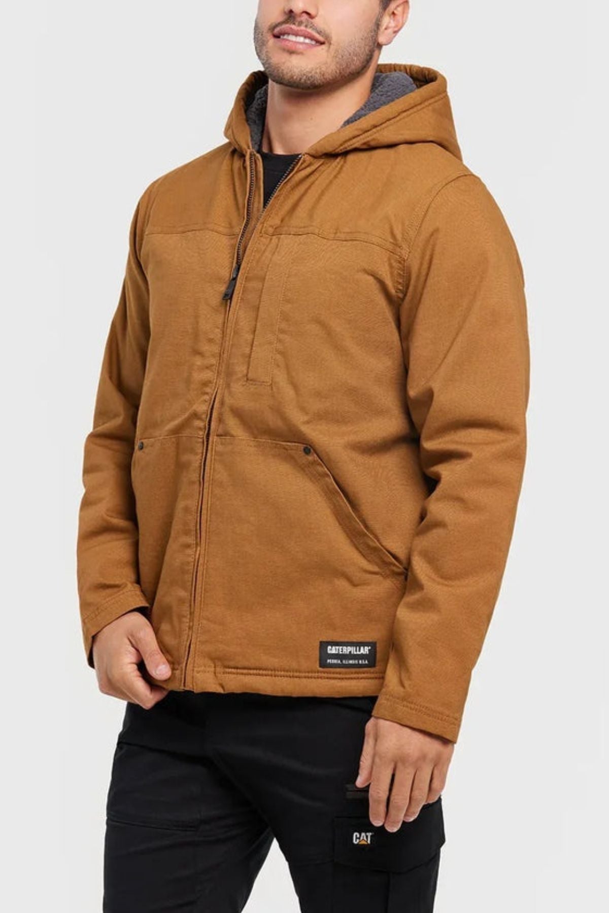 A man stands against a light background in black pants with a small CAT logo and a brown CAT Hooded Sherpa Lined Jacket featuring practical front pockets and a "Caterpillar" patch near the hem.