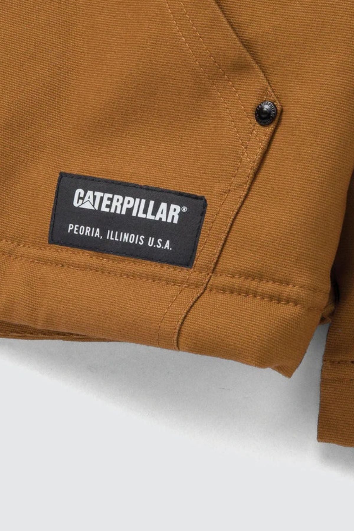 Close-up of a brown CAT Hooded Sherpa Lined Jacket with a black label reading "Caterpillar, Peoria, Illinois U.S.A.," visible stitching, and a black metal snap button.